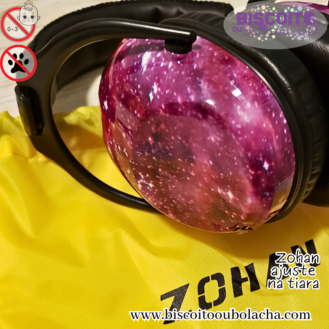 na imagem temos o abafador modelo ajuste na tiara da marca Zohan, com estampa galaxia na concha, sobre uma sacola de transporte de nylon na cor amarela com Zohan estampado em preto, e cordão preto no topo para fechar. Todas as conexões do abafador são de material plástico na cor preta, a capa acolchoada ao redor da tiara e a almofada da concha são em couro na cor preta. A estampa galáxia consiste em um fundo cor roxo, que vai variando a tonalidade pra roxo mais claro ou escuro, simulando o céu e diversos pontos irregulares brancos, que lembram estrelas no céu.